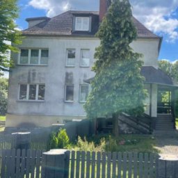 TWÓJ DACH Aleksander Mientki - Trzypiętrowy dom murowany z szarą elewacją, widoczny zza drewnianego płotu, z porośniętym mchem dachem i kominem.