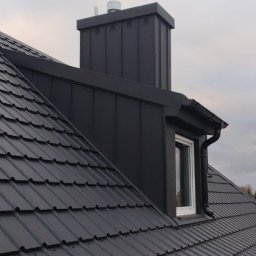TWÓJ DACH Aleksander Mientki - Czarny dach z dachówki ceramicznej, komin z blachy w kolorze dachu, okno dachowe z białą ramą i orynnowanie, widok z dołu na tle pochmurnego nieba.
