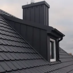 Czarny dach z dachówki ceramicznej, komin z blachy w kolorze dachu, okno dachowe z białą ramą i orynnowanie, widok z dołu na tle pochmurnego nieba.