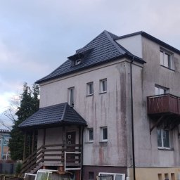 TWÓJ DACH Aleksander Mientki - Dwupiętrowy budynek z nowym, ciemnym dachem i wejściem z zadaszeniem, widoczne ślady zabrudzeń na elewacji, balkon z drewnianą balustradą i rusztowanie z boku budynku.