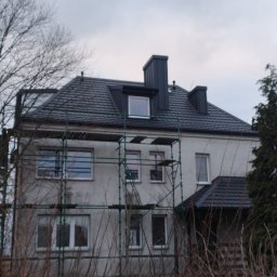 TWÓJ DACH Aleksander Mientki - Dwupiętrowy dom z ciemnoszarym dachem z dachówki, częściowo otoczony rusztowaniem, widoczny w pochmurny dzień.