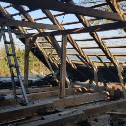 TWÓJ DACH Aleksander Mientki - Demontaż starej więźby dachowej z widoczną konstrukcją drewnianą, folią i drabiną opartą o krokwie, w tle zieleń drzew i fragment jeziora.