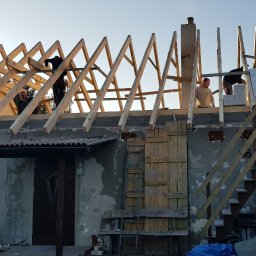 TWÓJ DACH Aleksander Mientki - Ekipa budowlana montuje drewnianą konstrukcję dachu na parterowym domu murowanym, widoczne krokwie i murłaty, w tle błękitne niebo.