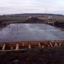Wylana płyta fundamentowa pod dom murowany w Dziemianach, z widocznym szalunkiem drewnianym i wystającymi prętami zbrojeniowymi, na tle krajobrazu z polem i lasem.
