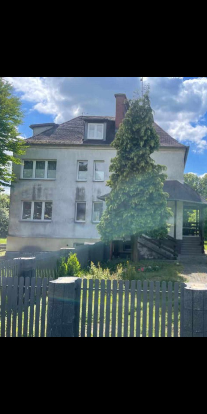 Trzypiętrowy dom murowany z szarą elewacją, widoczny zza drewnianego płotu, z porośniętym mchem dachem i kominem.