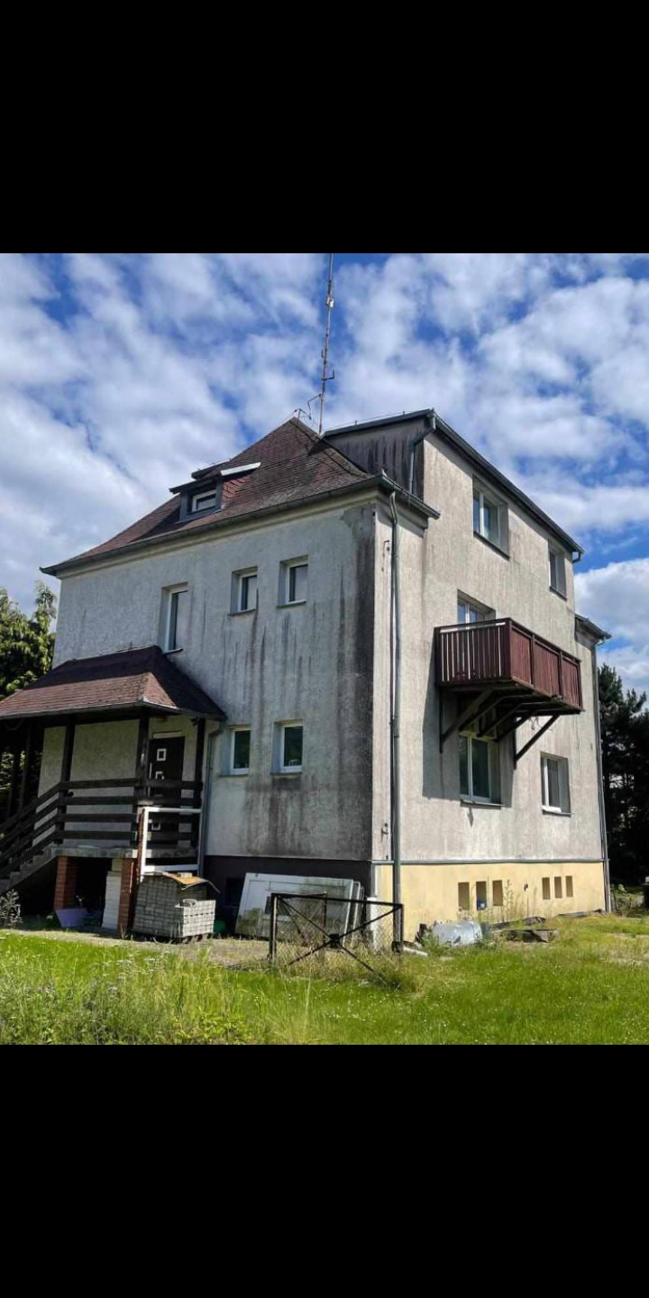 Dwukondygnacyjny dom z balkonem i werandą, widoczne ślady zabrudzeń na elewacji, antena na dachu, otoczenie zarośnięte trawą.