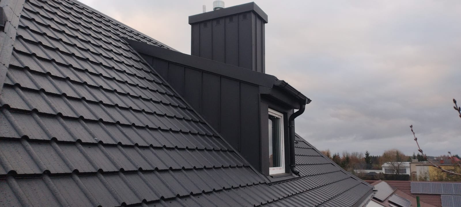 Czarny dach z dachówki ceramicznej, komin z blachy w kolorze dachu, okno dachowe z białą ramą i orynnowanie, widok z dołu na tle pochmurnego nieba.