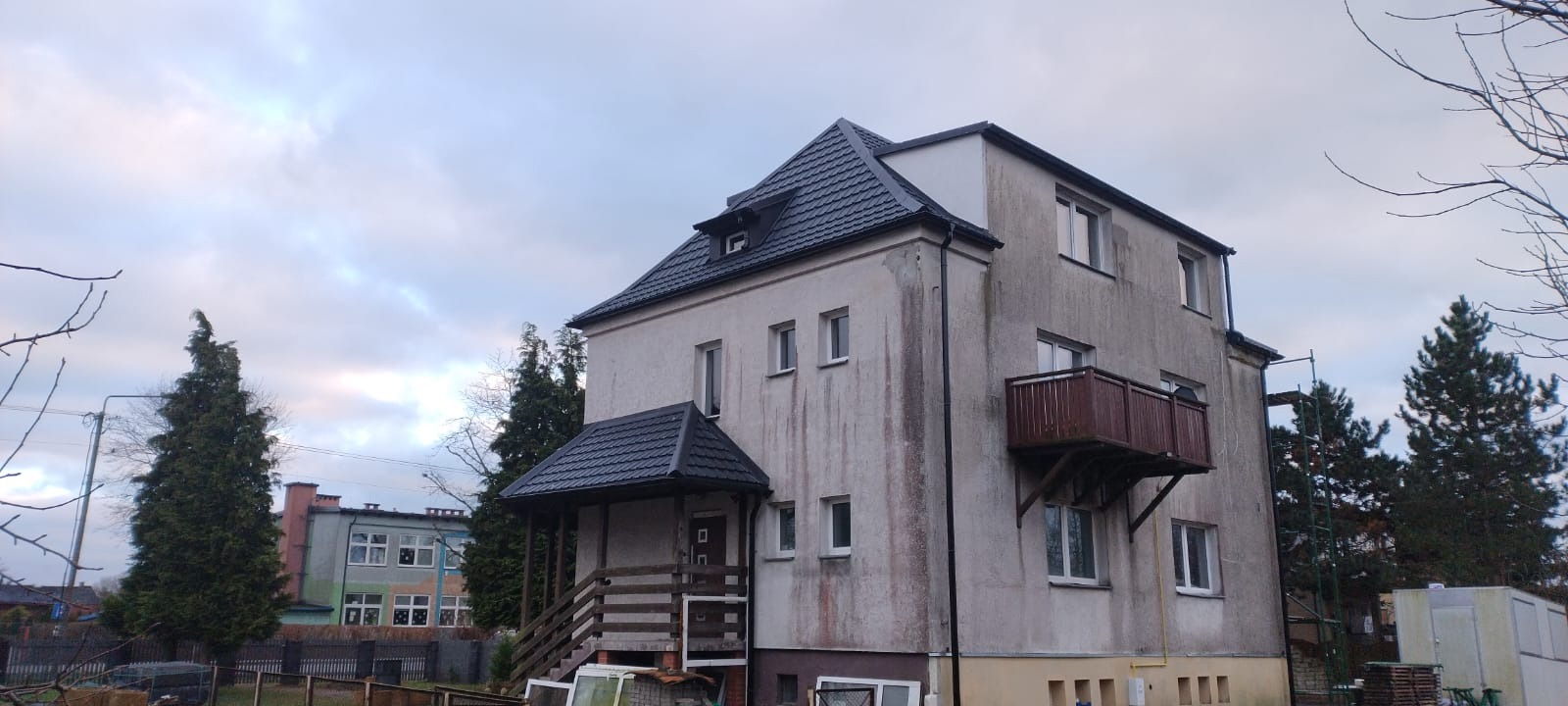 Dwupiętrowy budynek z nowym, ciemnym dachem i wejściem z zadaszeniem, widoczne ślady zabrudzeń na elewacji, balkon z drewnianą balustradą i rusztowanie z boku budynku.