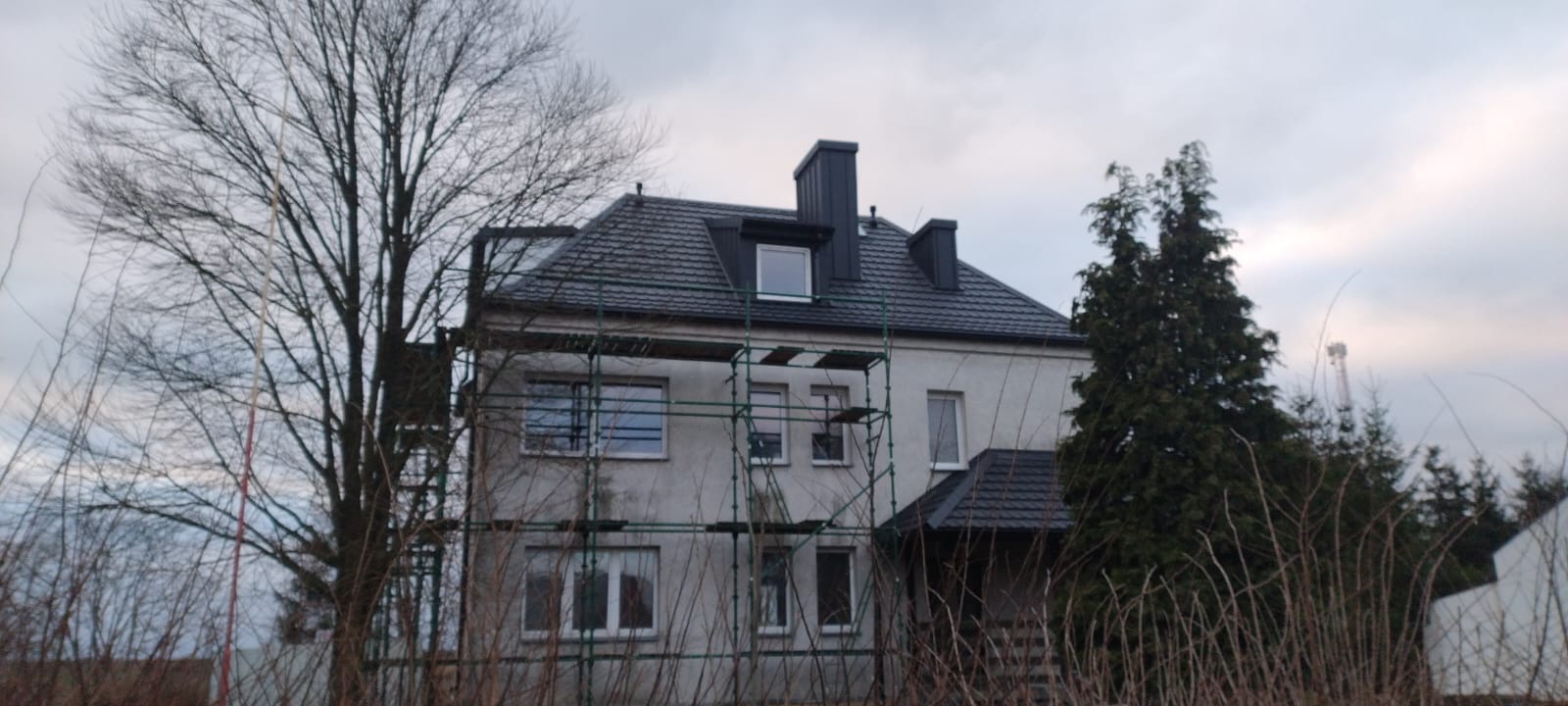Dwupiętrowy dom z ciemnoszarym dachem z dachówki, częściowo otoczony rusztowaniem, widoczny w pochmurny dzień.
