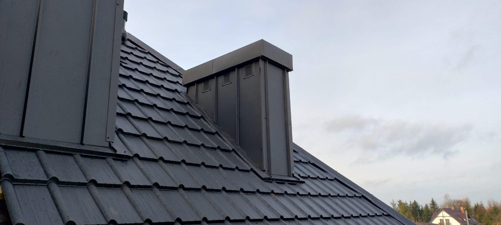 Ciemnoszary dach z dachówki ceramicznej z obróbkami blacharskimi komina i wentylacji, widok z dołu na tle pochmurnego nieba.