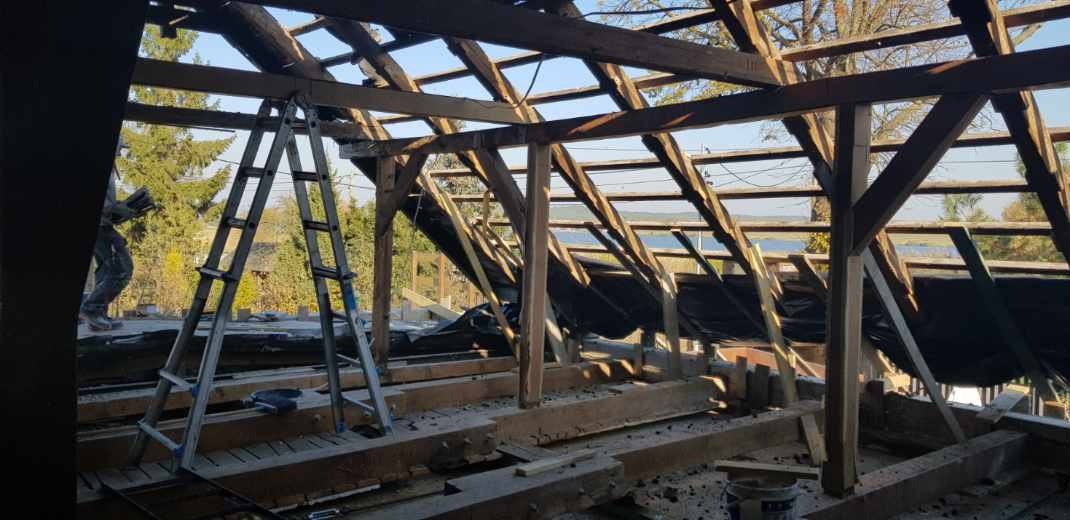 Demontaż starej więźby dachowej z widoczną konstrukcją drewnianą, folią i drabiną opartą o krokwie, w tle zieleń drzew i fragment jeziora.