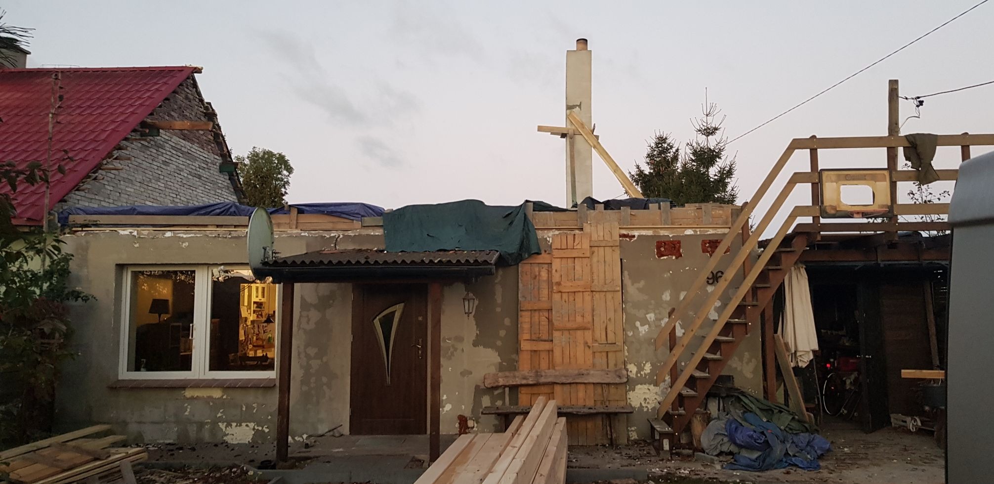 Dom w trakcie remontu dachu z widoczną konstrukcją drewnianą, zabezpieczony plandeką, z częściowo zdjętym czerwonym pokryciem i odsłoniętą cegłą, z elementami rusztowania i schodami na zewnątrz.