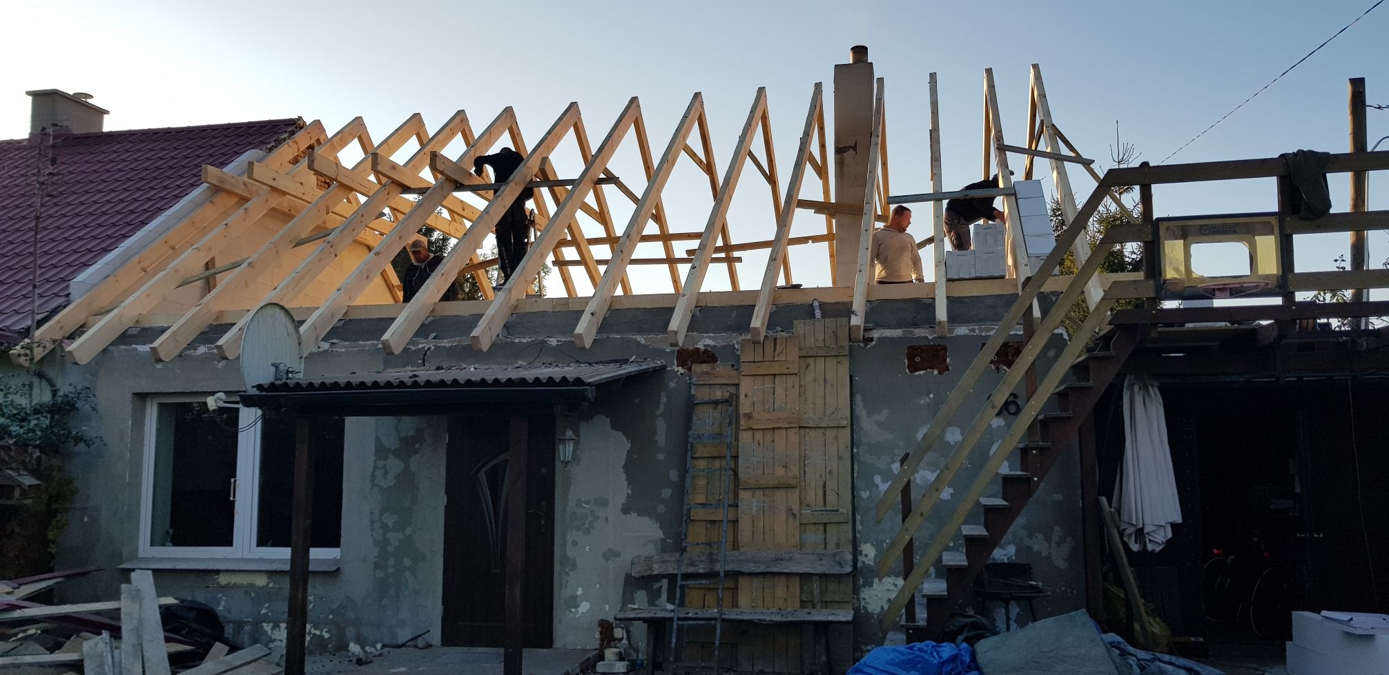 Ekipa budowlana montuje drewnianą konstrukcję dachu na parterowym domu murowanym, widoczne krokwie i murłaty, w tle błękitne niebo.