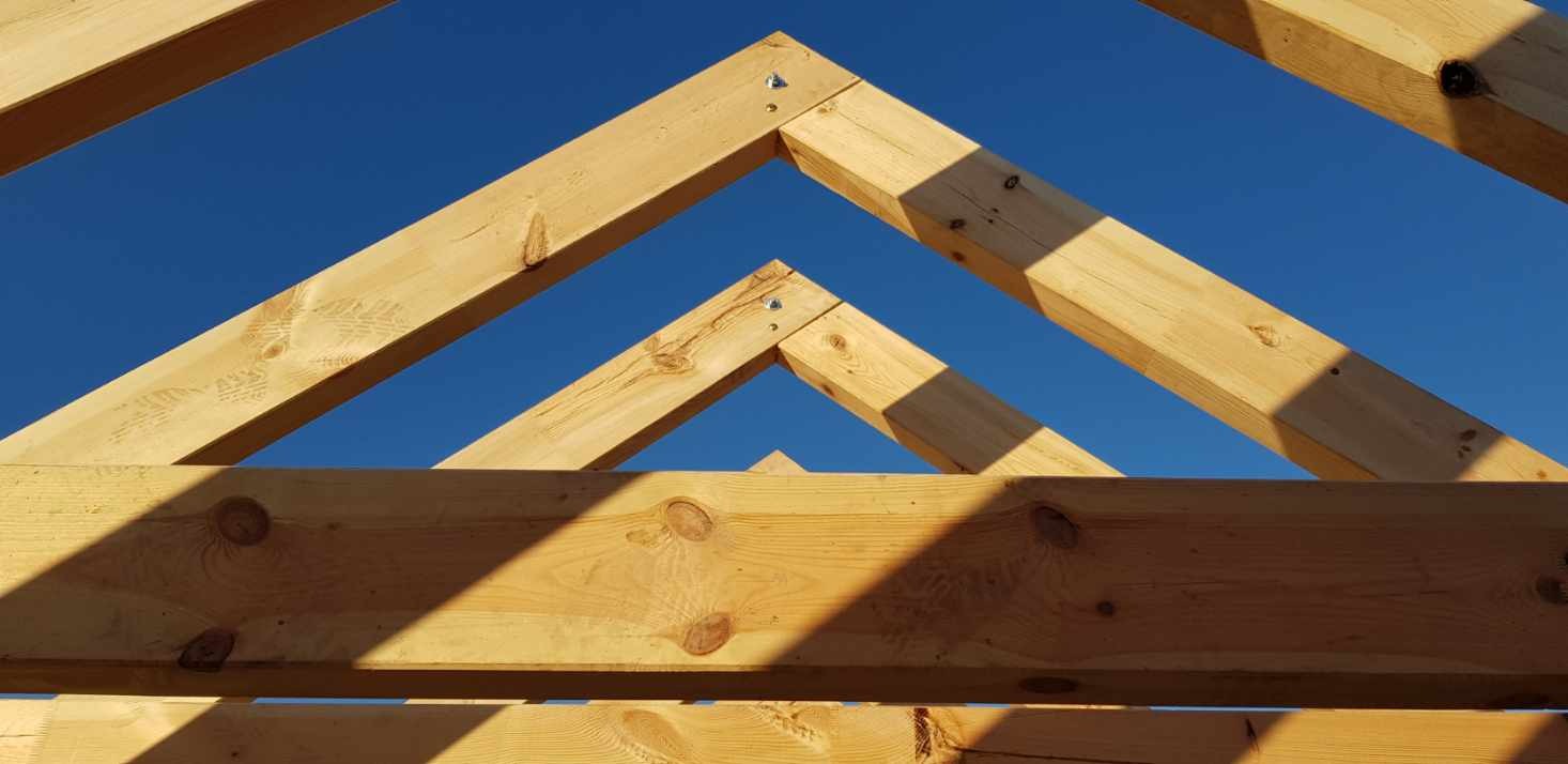 Fragment drewnianej konstrukcji więźby dachowej na tle błękitnego nieba, wykonanej w Dziemianach, z widocznymi detalami łączeń na śruby.