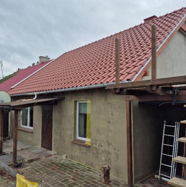 Dom z nowym, czerwonym dachem z dachówki ceramicznej, widoczne drewniane elementy konstrukcyjne i drabina, elewacja w trakcie wykańczania.
