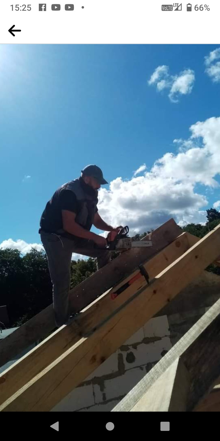 Pracownik w czapce i kamizelce tnie piłą łańcuchową drewniane elementy konstrukcji dachu, na tle błękitnego nieba z chmurami.