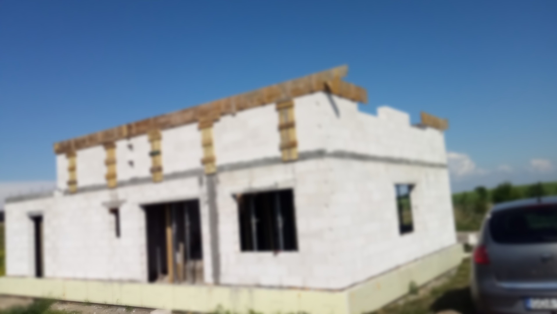 Surowy stan domu z białych bloczków, widoczne drewniane elementy konstrukcyjne dachu, nieostre tło z zielenią i fragmentem samochodu.