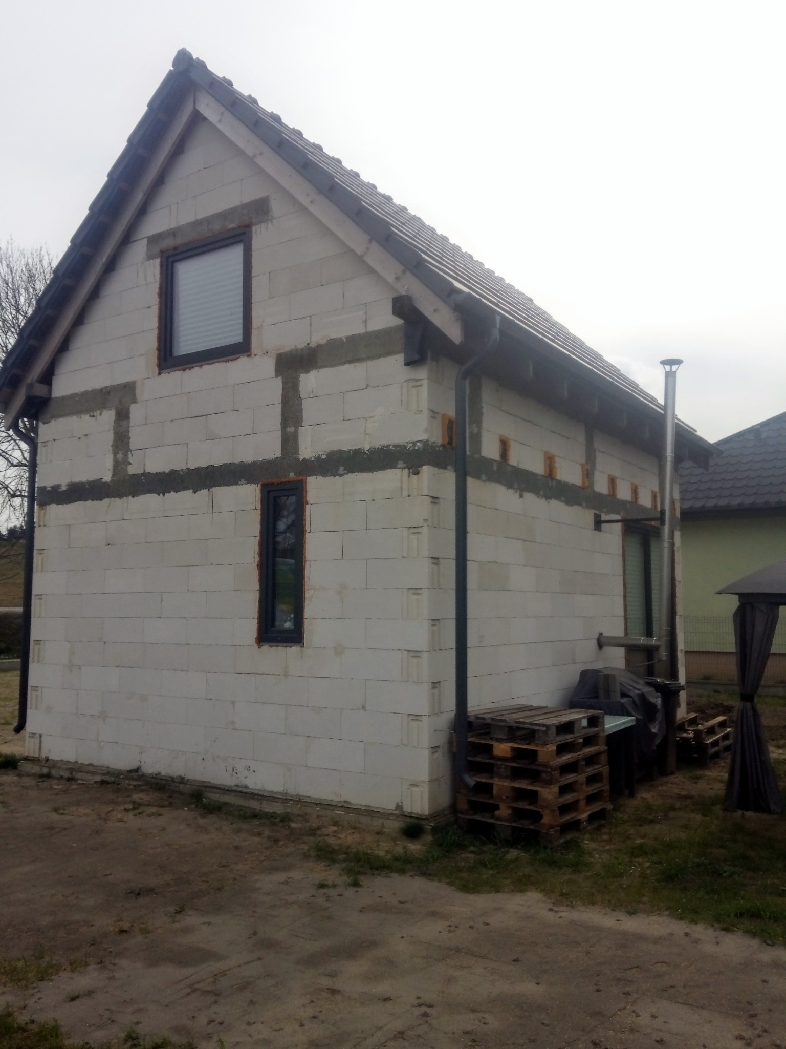 Surowy stan domu murowanego z białych bloczków, widoczne okna z szarą ramą, orynnowanie i palety obok budynku.