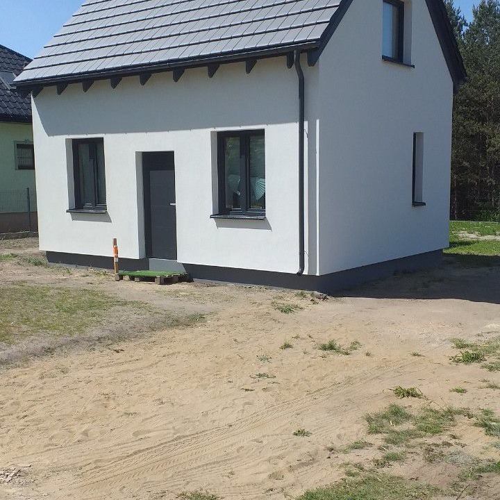 Nowo wybudowany, jednopiętrowy dom murowany z szarym dachem i czarnymi ramami okiennymi, widoczny z boku, z tymczasowym wejściem na palecie.