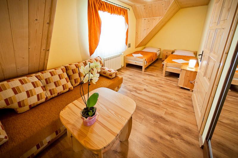 Pokój w pensjonacie na poddaszu z drewnianą boazerią, dwoma łóżkami, kanapą z poduszkami w kratę i stolikiem z orchideą w doniczce.