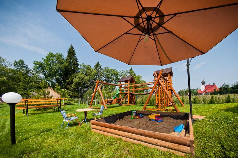 Plac zabaw z piaskownicą, huśtawkami i zjeżdżalnią na zielonym trawniku, częściowo zacieniony brązowym parasolem, z widokiem na budynek z czerwoną dachówką w oddali.