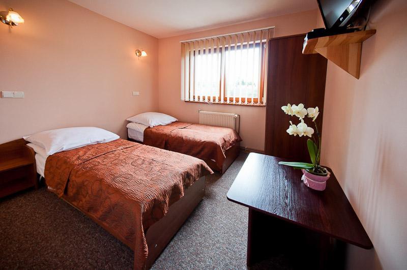 Pokój dwuosobowy z brązowymi narzutami, białymi poduszkami, ciemnym drewnianym stolikiem z orchideą i telewizorem na ścianie.