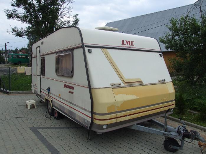 Używana przyczepa kempingowa LMC 490P Dominat na brukowanym podwórku, widoczny hak holowniczy i biały stołek obok koła, w tle budynek gospodarczy i zielony autobus.