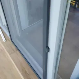 Zamknięta moskitiera w oknie przesuwnym, widok z bliska na mechanizm blokujący, jasne panele podłogowe.