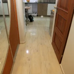 Połyskująca, kremowa podłoga z płytek imitujących drewno, ułożona w długim korytarzu z widokiem na kuchnię. Częściowo widoczna szafa z lustrem i drewniane drzwi.