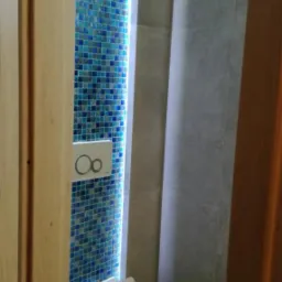 Nowoczesna łazienka z mozaiką w odcieniach błękitu, podświetleniem LED i minimalistyczną toaletą z przyciskiem spłukującym w kształcie dwóch kółek.