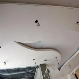 Biały sufit z dekoracyjnym elementem z płyty karton gips i otworami na oświetlenie punktowe, w trakcie montażu. Widoczny fragment drabiny i zabezpieczenia podłogi.