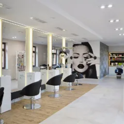 Nowoczesne wnętrze salonu fryzjerskiego z stanowiskami do stylizacji, czarnymi fotelami, dużym muralem kobiety i oświetleniem LED.