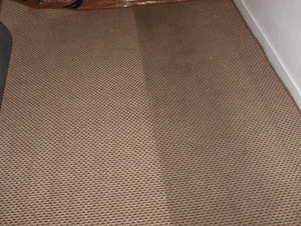 Beżowy dywan o wyraźnej fakturze typu plaster miodu, widoczne ślady po czyszczeniu, zarys mebla w lewym górnym rogu.