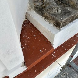 Z.U.H. MATEX-BUD Mateusz Wacławski - Fragment elewacji budynku z detalami architektonicznymi, w tym cokół z rzeźbionymi stopami figury, oraz nowo zamontowana, brązowa blacha okapowa, zabezpieczająca przed wodą, widoczna z bliska...