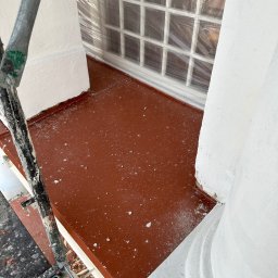 Z.U.H. MATEX-BUD Mateusz Wacławski - Balkon w trakcie renowacji: widok z rusztowania na pomalowaną na brązowo podłogę, zabezpieczone folią okno i świeżo pomalowane na biało filary.