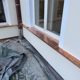 Z.U.H. MATEX-BUD Mateusz Wacławski - Montaż miedzianych obróbek blacharskich wokół okien na elewacji budynku, widoczne narzędzia i materiały budowlane na plandece ochronnej.