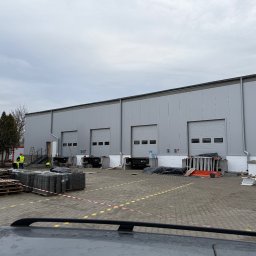 Z.U.H. MATEX-BUD Mateusz Wacławski - Budowa hali z szarej płyty warstwowej, widoczni robotnicy w odblaskowych kamizelkach, palety z kostką brukową i materiały budowlane na placu budowy, pochmurne niebo.