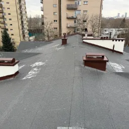 Widok z góry na płaski dach pokryty papą, z widocznymi kominami w brązowo-białej kolorystyce, delikatnie przyprószony śniegiem, z budynkami mieszkalnymi i panoramą miasta w tle.