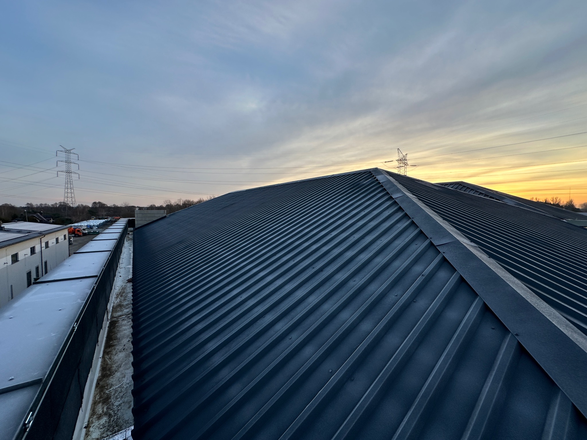 Widok z góry na nowo położone, ciemne, blaszane pokrycie dachu hali przemysłowej, z widoczną linią kalenicy i słupami wysokiego napięcia na horyzoncie, sfotografowane o zmierzchu.