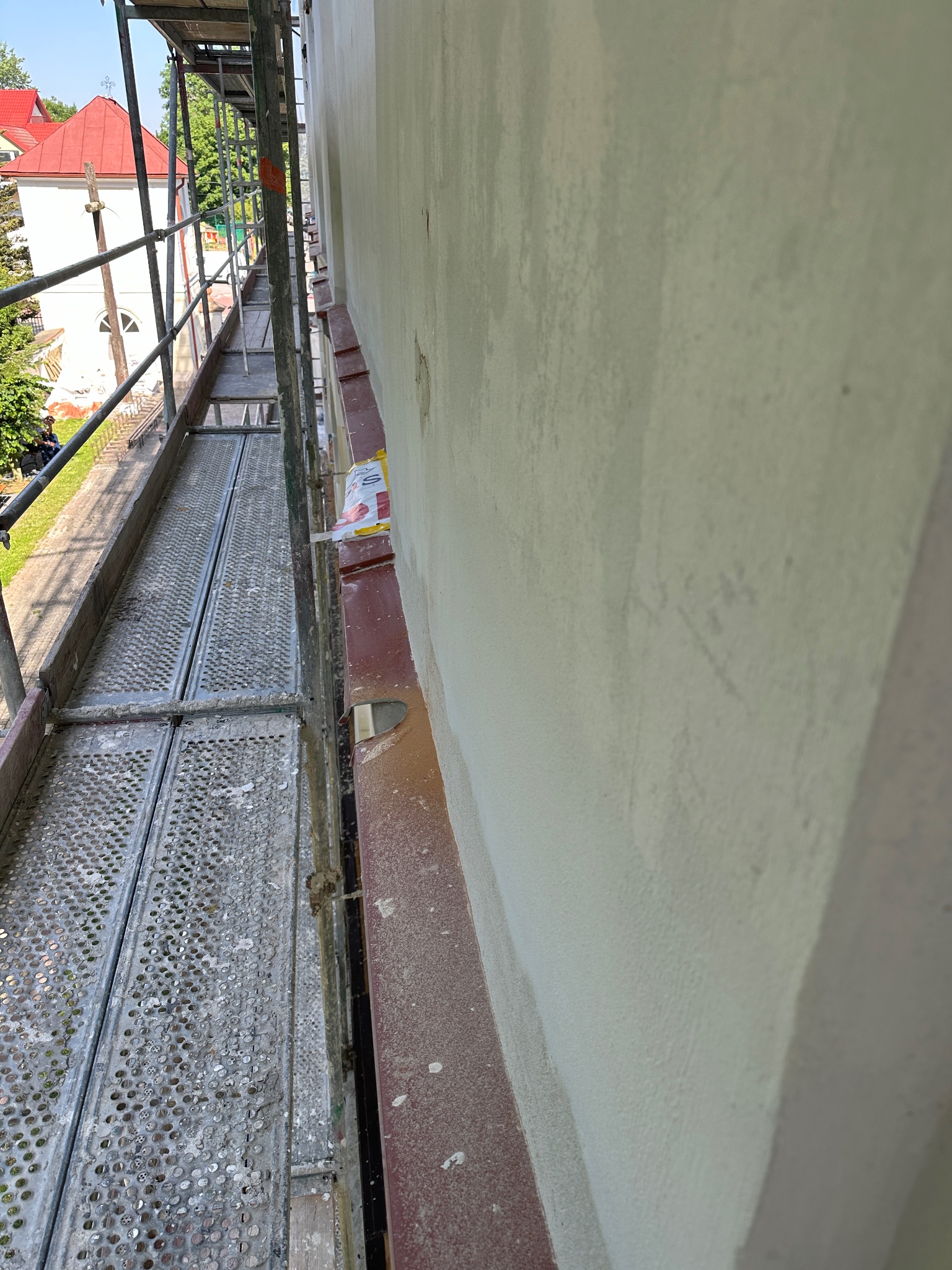 Widok ze stalowego rusztowania na ścianę budynku w trakcie prac wykończeniowych; widoczna blacha okapowa i fragment budynku sakralnego w tle.