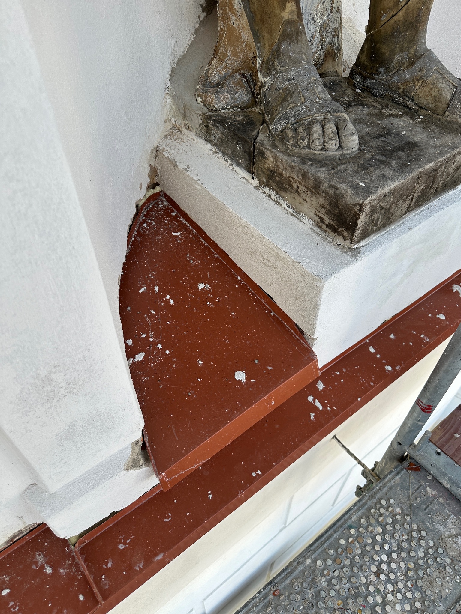 Fragment elewacji budynku z detalami architektonicznymi, w tym cokół z rzeźbionymi stopami figury, oraz nowo zamontowana, brązowa blacha okapowa, zabezpieczająca przed wodą, widoczna z bliska...