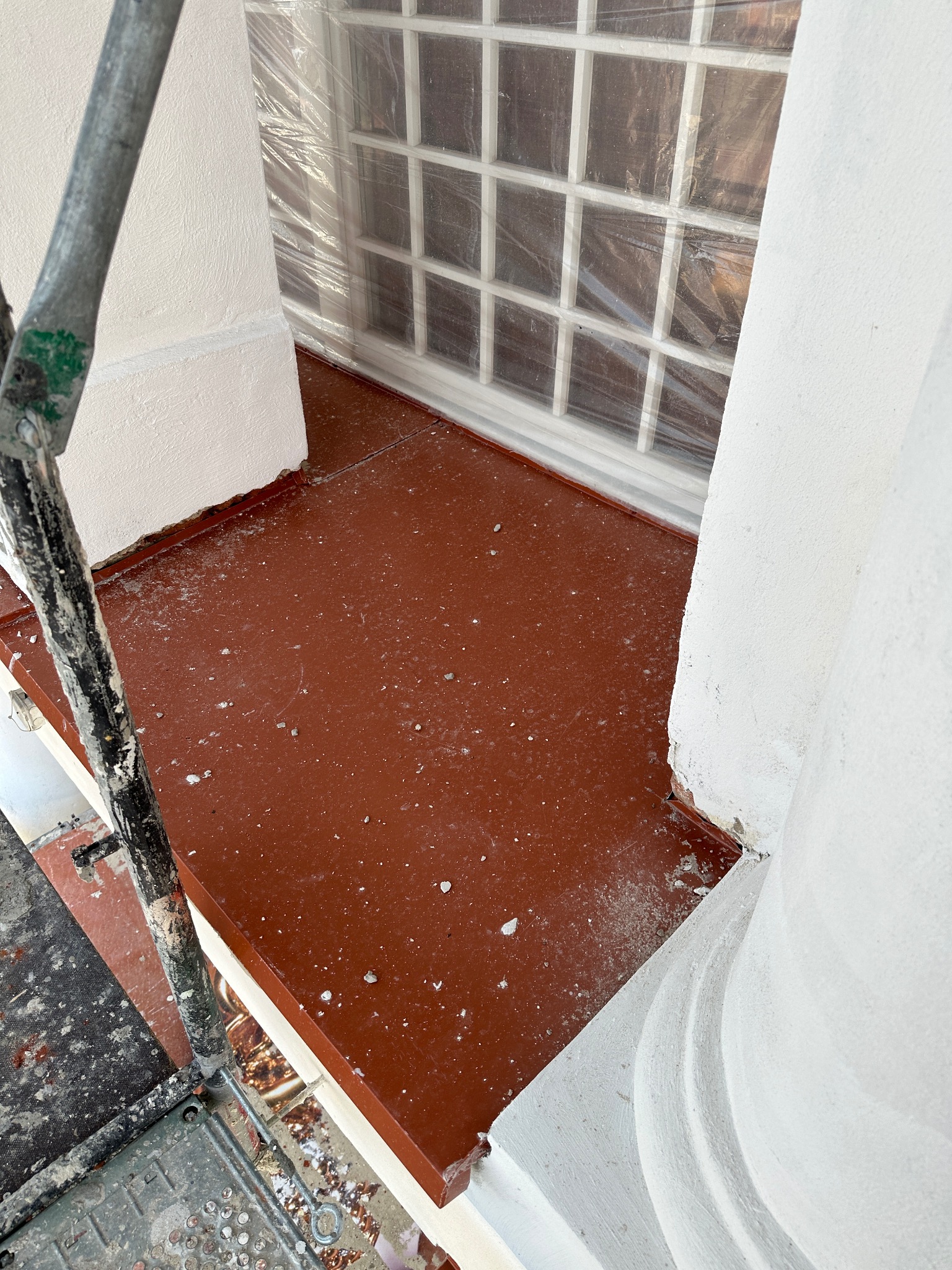 Balkon w trakcie renowacji: widok z rusztowania na pomalowaną na brązowo podłogę, zabezpieczone folią okno i świeżo pomalowane na biało filary.