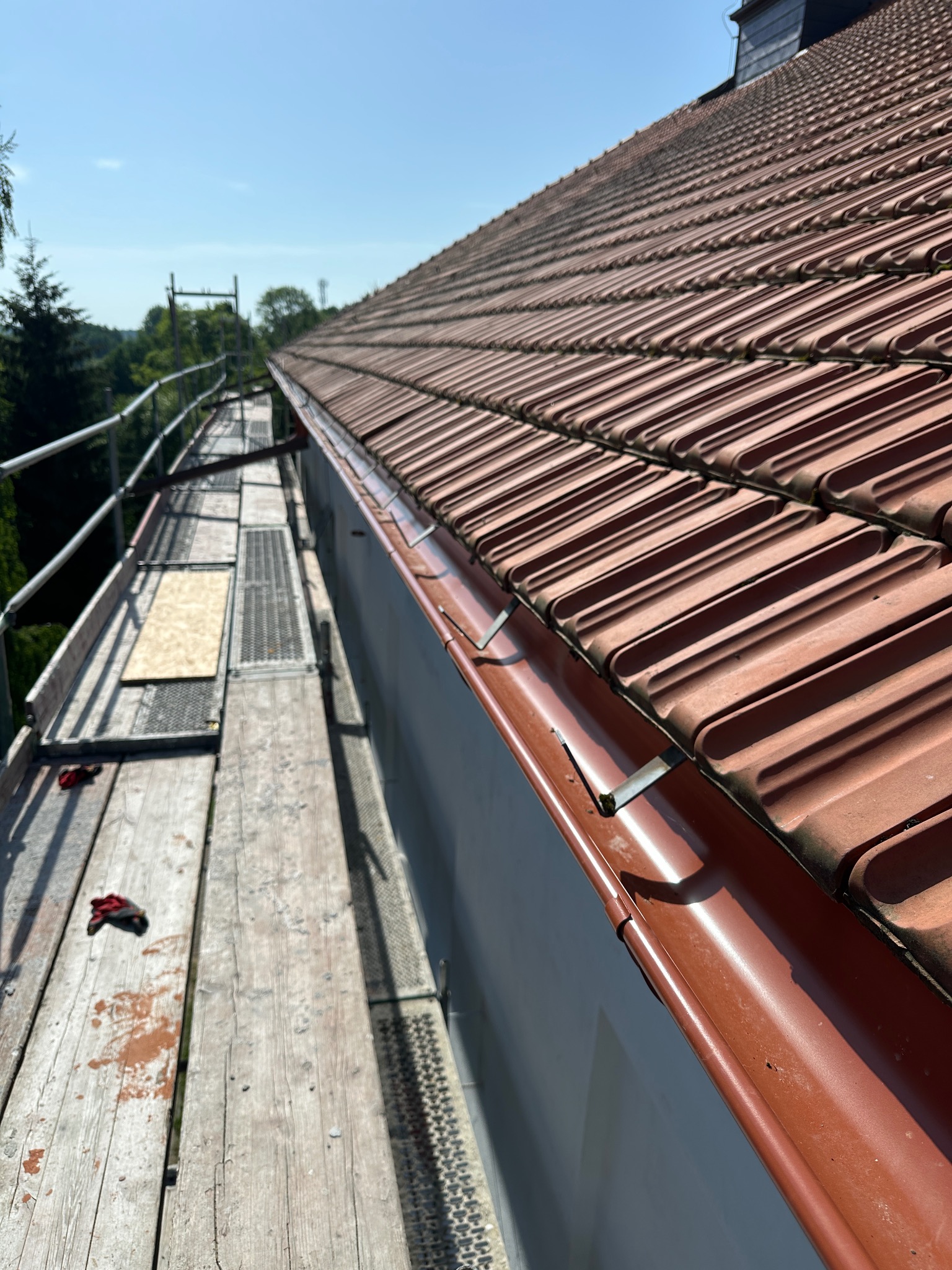 Czerwona dachówka ceramiczna na skośnym dachu, widoczna z rusztowania z metalowymi rynnami w słoneczny dzień.