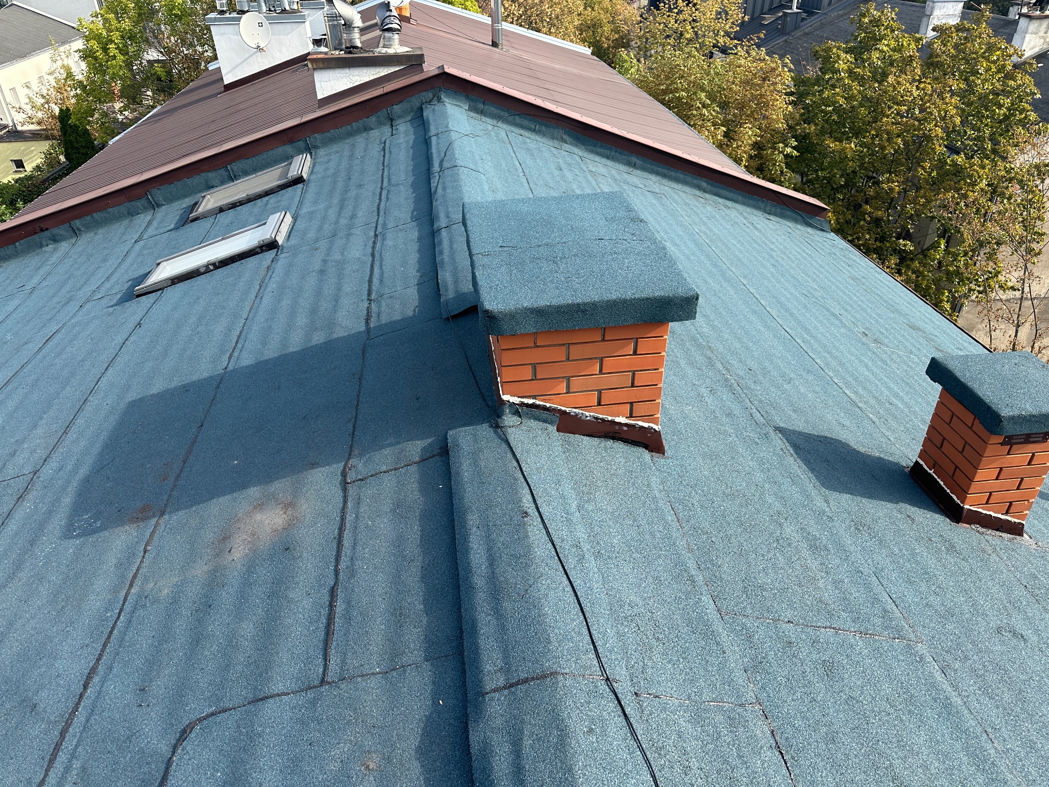 Niebieski dach pokryty papą z dwoma ceglanymi kominami i oknami dachowymi, widok z góry na tle budynków i drzew