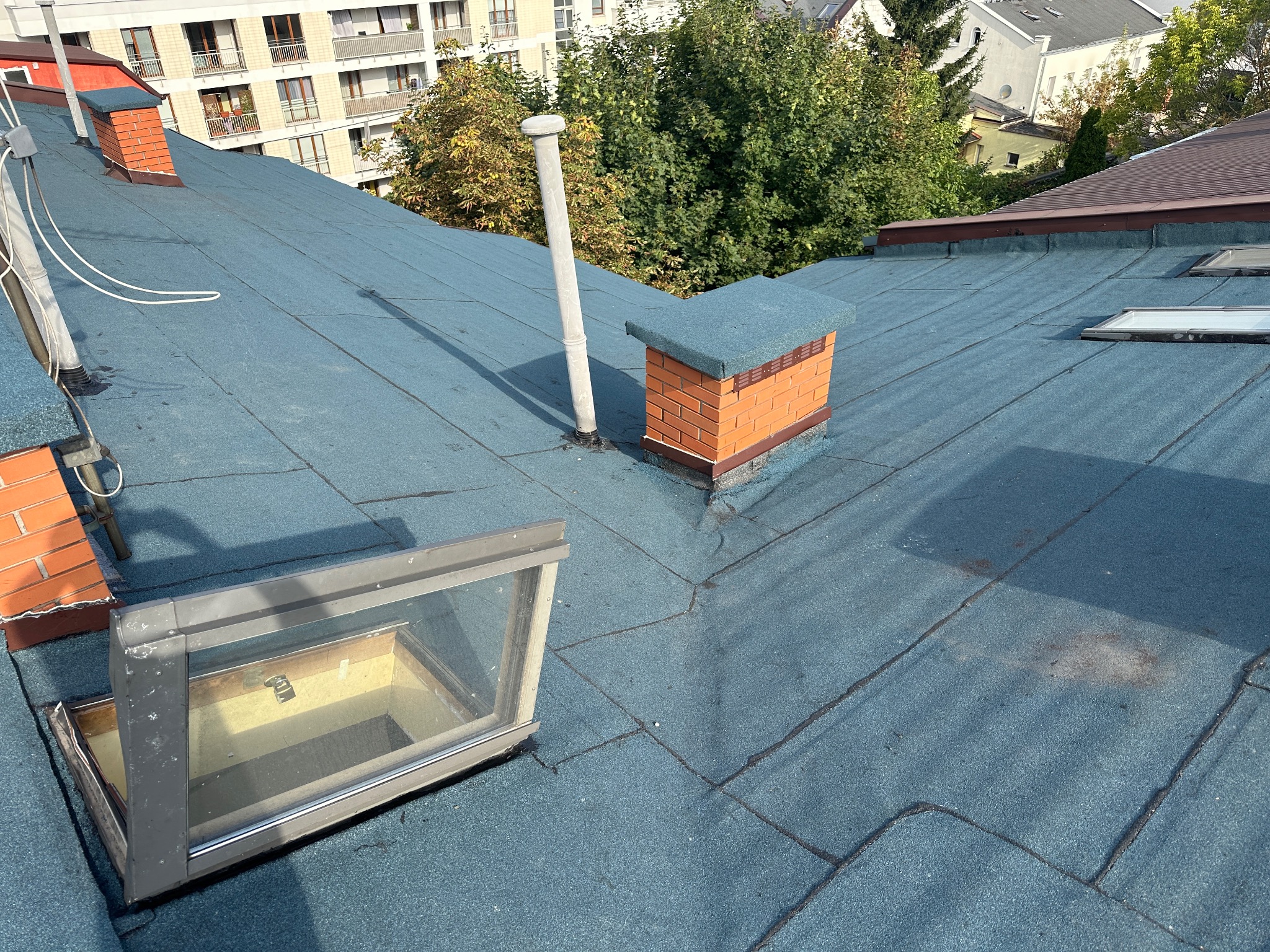 Niebieski dach pokryty papą z widocznym kominem z cegły, rurą wentylacyjną i otwartym oknem dachowym, na tle drzew i budynków.