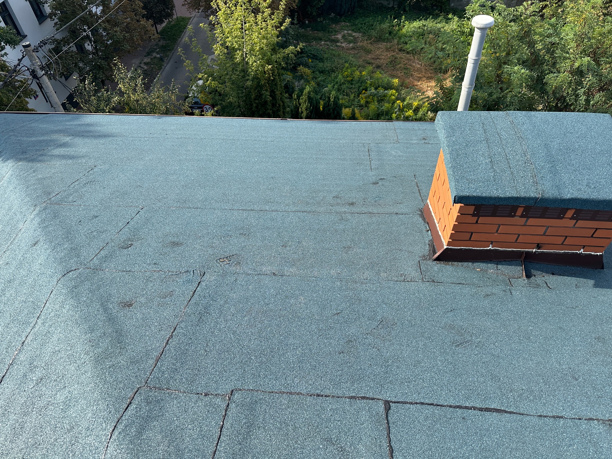 Widok z góry na płaski dach pokryty papą termozgrzewalną w kolorze niebiesko-szarym, z widocznym kominem obłożonym cegłą i fragmentem wentylacji, otoczony zielenią drzew.