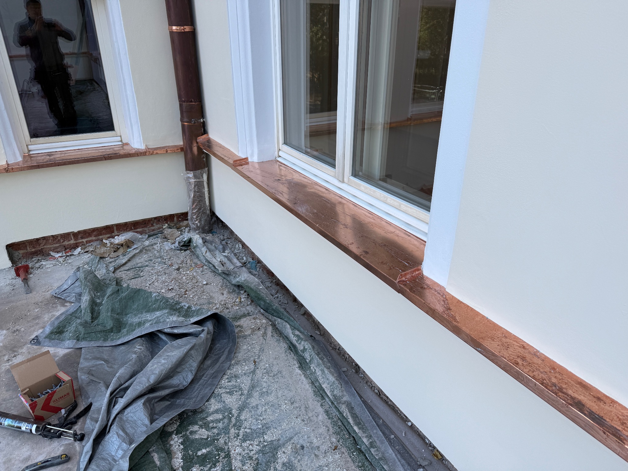 Montaż miedzianych obróbek blacharskich wokół okien na elewacji budynku, widoczne narzędzia i materiały budowlane na plandece ochronnej.