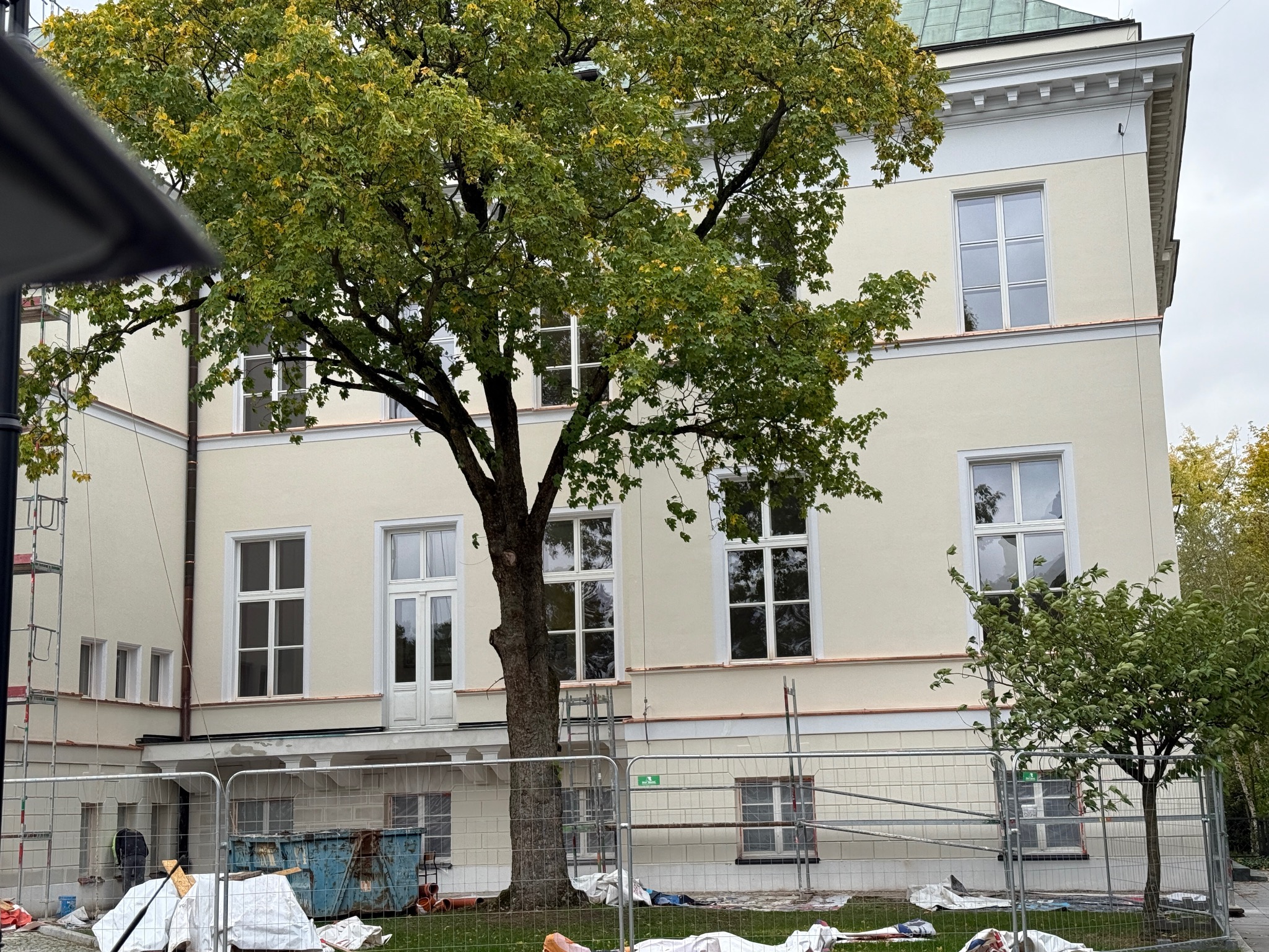 Budynek w trakcie renowacji elewacji, widoczne rusztowania, zabezpieczenia i kontener budowlany, ujęcie zza ogrodzenia placu budowy.