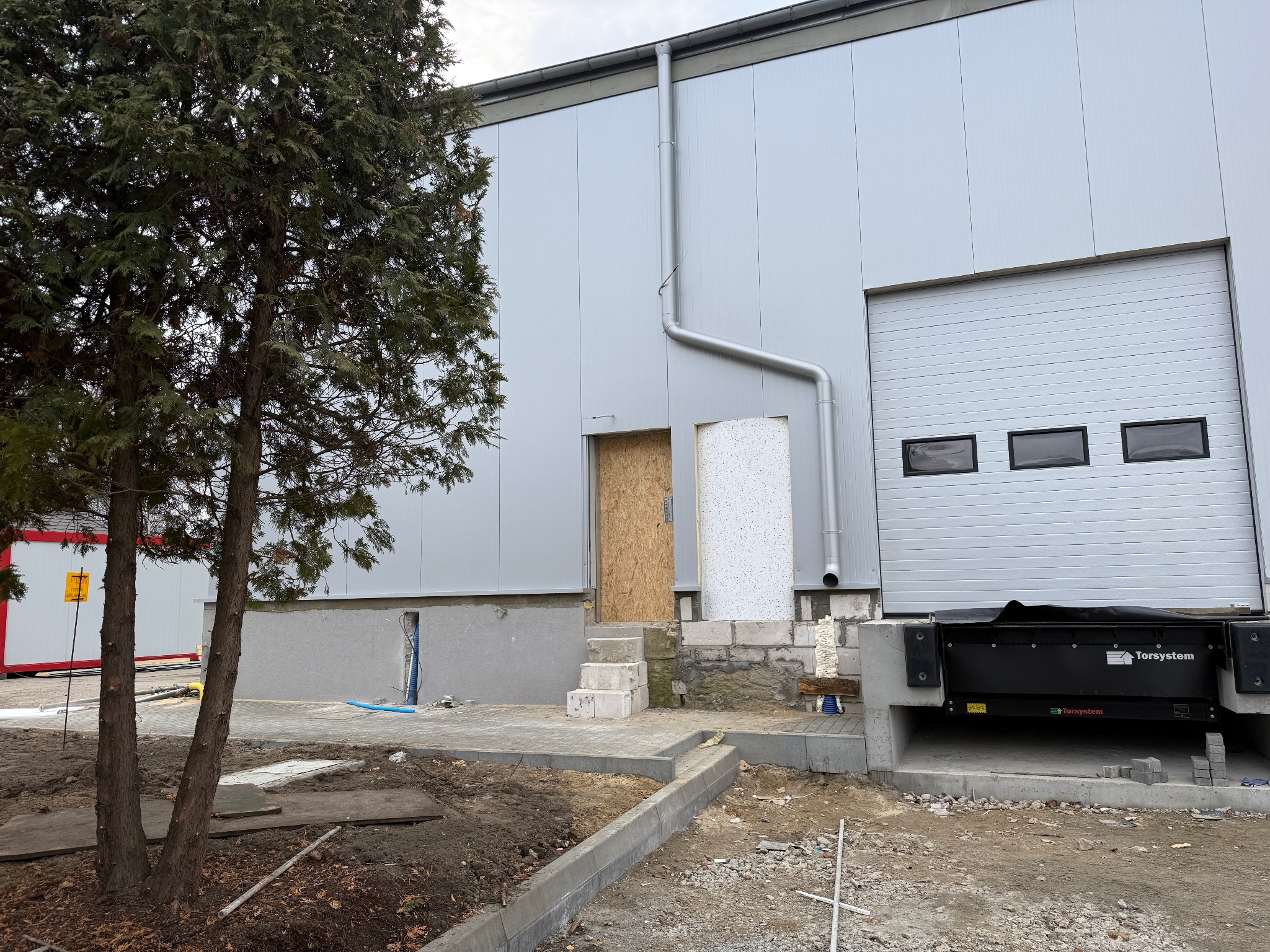 Szara hala z panelowych płyt, z widocznymi drzwiami wejściowymi z płyt OSB i bramą segmentową z trzema oknami, rynna spustowa po prawej stronie, teren wokół w trakcie prac wykończeniowych.
