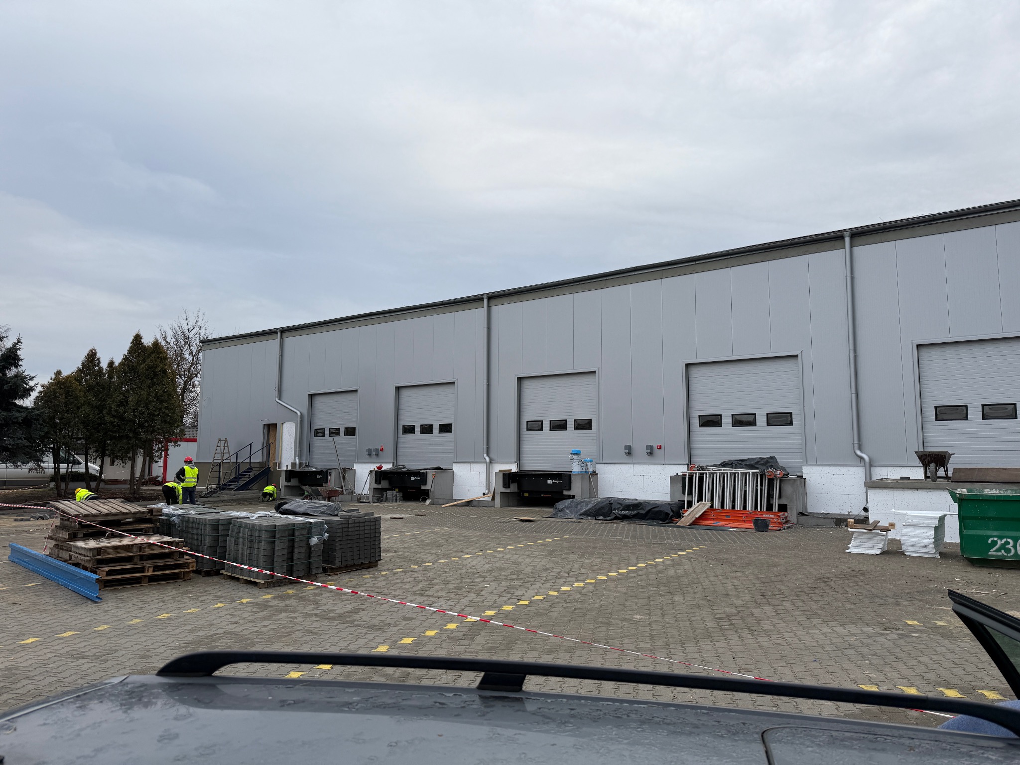 Budowa hali z szarej płyty warstwowej, widoczni robotnicy w odblaskowych kamizelkach, palety z kostką brukową i materiały budowlane na placu budowy, pochmurne niebo.